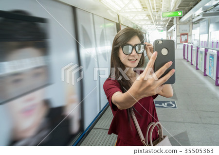 Asian woman passenger with casual suit taking selfie by the smart mobile phone in the BTS Skytrain rails or MRT subway for travel in the big city, lifestyle and transportation concept 35505363