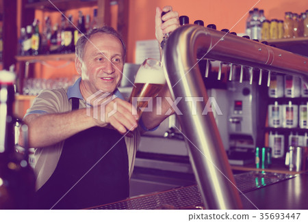 Barman is pouring unbottled beer with foam for client in the bar. Barman is pouring unbottled beer with foam for client in the bar. 35693447