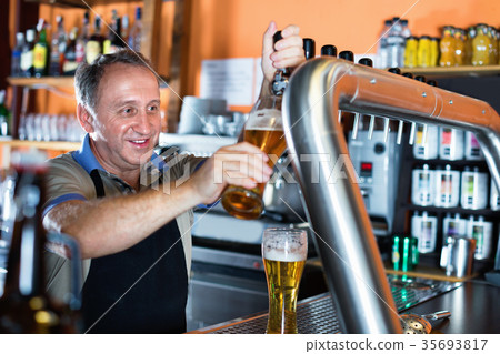 Barman is pouring unbottled beer with foam for client in the bar. 35693817