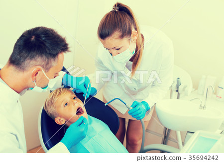 Stomatologist with girl assistant are diagnosticating to young patient which is sitting 35694242