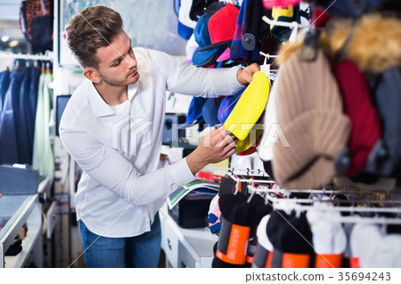 Smiling positive guy deciding on warm hat in sports store 35694243