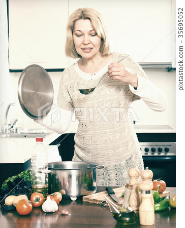 Happy mature housewife with ladle cooking soup in pan in kit Happy mature housewife with ladle cooking soup in pan in kit 35695052