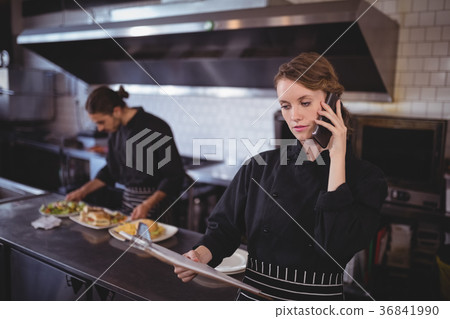 Young waitress talking on smartphone while waiter preparing food in commercial kitchen 36841990