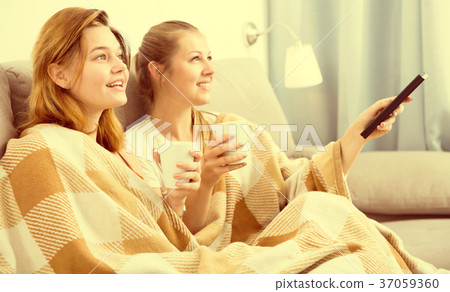 Two young women in plaide are watching TV and warming with tea Two young women in plaide are watching TV and warming with tea 37059360