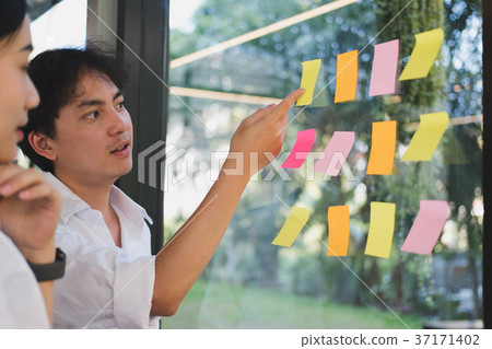 man & woman discussing creative idea with adhesive notes on glass wall at workplace. Sticky note paper reminder schedule at office. business, brainstorming, creativity concept man & woman discussing creative idea with adhesive notes on glass wall at workplace. Sticky note paper reminder schedule at office. business, brainstorming, creativity concept 37171402