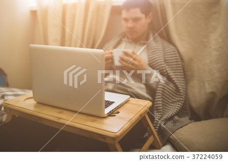 Young man laying in bad with laptop and cup of tea Young man laying in bad with laptop and cup of tea 37224059