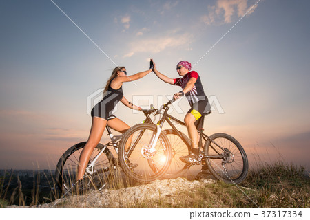 Cyclist couple with mountain bikes on the hill at sunset Cyclist couple with mountain bikes on the hill at sunset 37317334