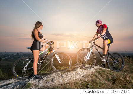Cyclist couple with mountain bikes on the hill at sunset Cyclist couple with mountain bikes on the hill at sunset 37317335
