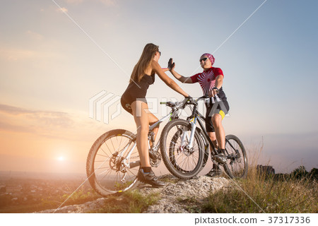 Cyclist couple with mountain bikes on the hill at sunset Cyclist couple with mountain bikes on the hill at sunset 37317336