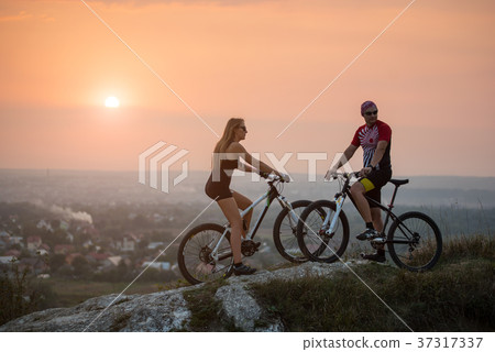 Cyclist couple with mountain bikes on the hill at sunset Cyclist couple with mountain bikes on the hill at sunset 37317337