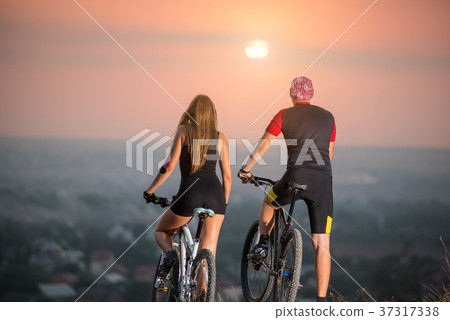 Couple bicyclist with mountain bikes on the hill at sunset Couple bicyclist with mountain bikes on the hill at sunset 37317338