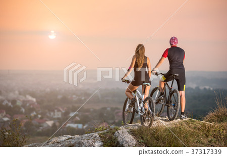 Cyclist couple with mountain bikes on the hill at sunset Cyclist couple with mountain bikes on the hill at sunset 37317339