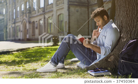Multiracial young guy sitting under tree, reading 37450364