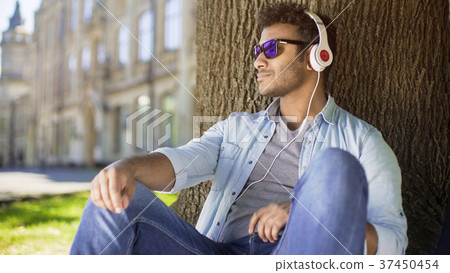 Multiracial guy leaning against tree in headphones Multiracial guy leaning against tree in headphones 37450454
