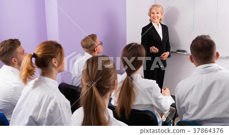 Group of medical students attentively listening to lecture of female teacher in classroom Group of medical students attentively listening to lecture of female teacher in classroom 37864376