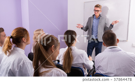 Young male lecturer answering near whiteboard in auditorium Young male lecturer answering near whiteboard in auditorium 37864478