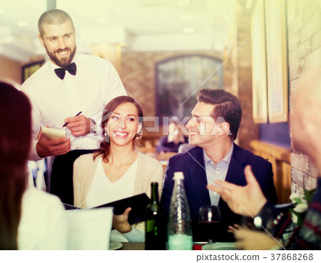 Waiter with notebook taking order from friendly company indoors Waiter with notebook taking order from friendly company indoors 37868268