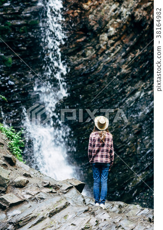 Girl in hat stands on rock and looks at waterfall Girl in hat stands on rock and looks at waterfall 38164982