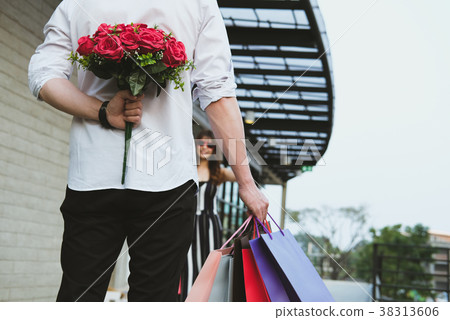 man holding shopping bag & bouquet of red roses behind his back for woman. boyfriend surprise girlfriend with flower in valentine's day. celebrate anniversary, love, relationship concept 38313606