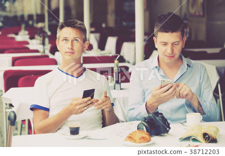 Two men sitting with coffee and looking at mobiles in cafe Two men sitting with coffee and looking at mobiles in cafe 38712203