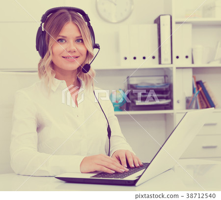 Young call center employee in headphones is sitting near laptop in the office Young call center employee in headphones is sitting near laptop in the office 38712540