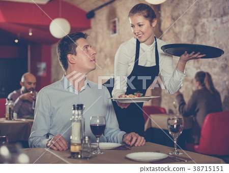 Polite waitress serving ordered dishes to smiling man at restaur 38715151