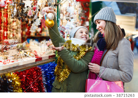 Smiling female and her daughter are choosing decorations for Christmas tree in the market outdoor. 38715263