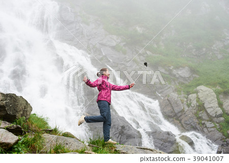 Smiling young female making selfie posing on rock in front of magnificent Balea waterfall in Romania Smiling young female making selfie posing on rock in front of magnificent Balea waterfall in Romania 39041007