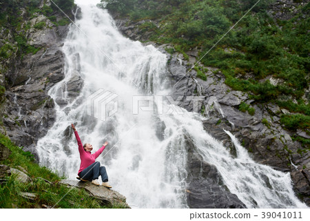 Young woman sits with hands up in front waterfall Balea Young woman sits with hands up in front waterfall Balea 39041011
