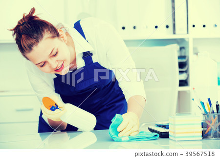 Cheerful female cleaner is cleaning dust from the desk Cheerful female cleaner is cleaning dust from the desk 39567518