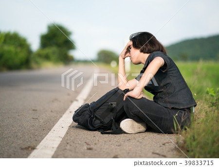 Woman sit with backpack hitchhiking along a road in countryside Woman sit with backpack hitchhiking along a road in countryside 39833715