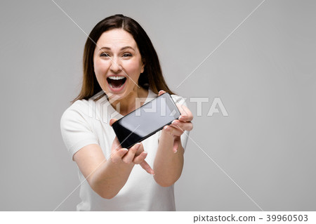 Emotional smiling happy plus size model standing in studio looking in camera offering mobile phone 39960503