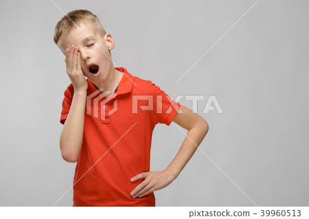 Portrait of blonde caucasian sad little boy in orange t-shirt wanting to sleep yawning on gray 39960513