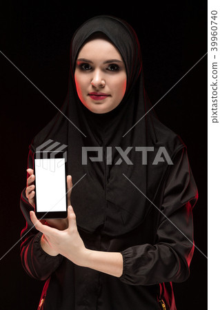 Portrait of beautiful smart young muslim woman wearing black hijab advertising mobile phone in her Portrait of beautiful smart young muslim woman wearing black hijab advertising mobile phone in her 39960740