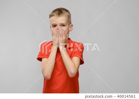 Portrait of blonde caucasian sad little boy in orange t-shirt closing his mouth with hands on gray 40014561
