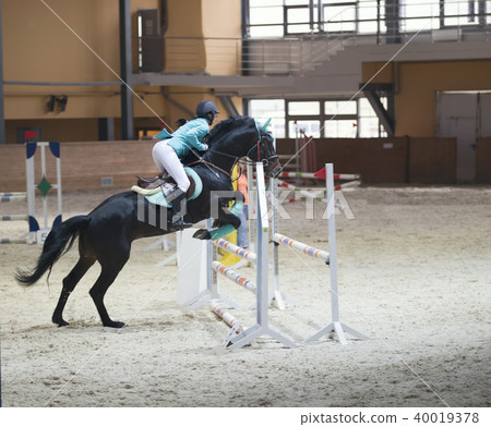 Young woman on the black stallion jumping over hurdle at show jumping competition 40019378