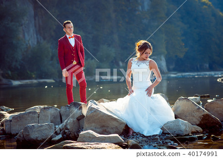 Charming red head bride in long dress is sitting on the stone near the river while groom in red suit 40174991
