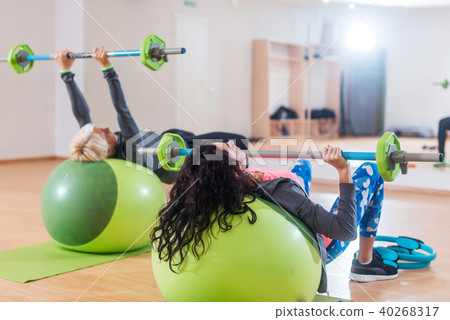 Back view of two women lifting barbell lying on stability ball while exercising in gym Back view of two women lifting barbell lying on stability ball while exercising in gym 40268317