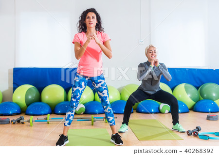 Two women in sportswear doing bodyweight squats while training indoors at sports hall 40268392