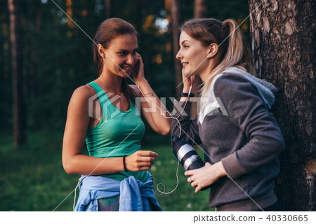 Two female runners relaxing after workout standing near the tree talking in park 40330665