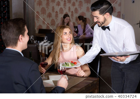 Polite waiter bringing ordered dishes to smiling couple at restaurant Polite waiter bringing ordered dishes to smiling couple at restaurant 40334724