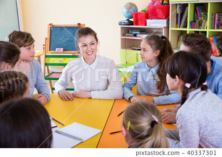 Friendly female teacher talking to children, sitting together around desk Friendly female teacher talking to children, sitting together around desk 40337301