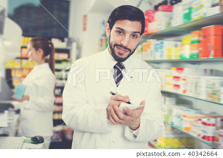 Male specialist is attentively stocktaking medicines with notebook near shelves in pharmacy. Male specialist is attentively stocktaking medicines with notebook near shelves in pharmacy. 40342664