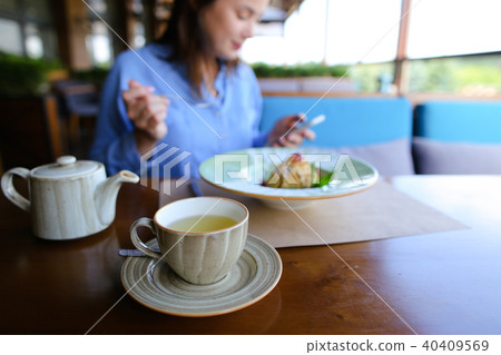 Close up cup of tea, kettle and female person eating dessert in background at cafe, using smartphone Close up cup of tea, kettle and female person eating dessert in background at cafe, using smartphone 40409569