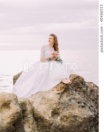 The bride in the long wedding dress is sitting on the cliff and watching the sunset at the The bride in the long wedding dress is sitting on the cliff and watching the sunset at the 40498553