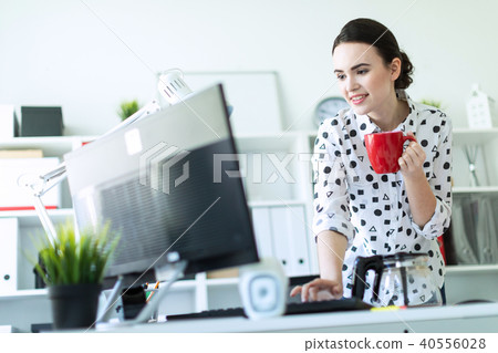 A young girl is standing in the office near the table, holding a red mug in her hand and typing on 40556028