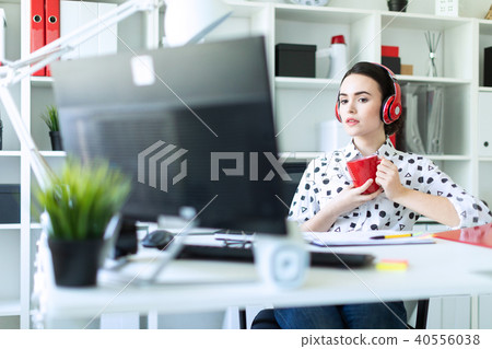 A young girl sits in headphones at a table in the office, holds a red cup in her hands and looks at 40556038