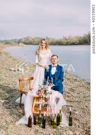 The bride is standing behing the sitting groom at the table placed on the coast. 40559903