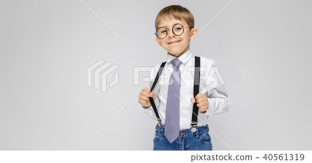 A charming boy in a white shirt, suspenders, a tie and light jeans stands on a gray background. the 40561319