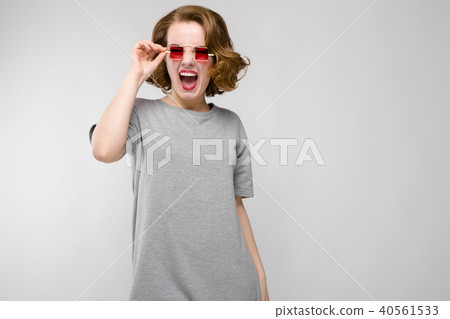 Charming young girl in a gray T-shirt on a gray background. Girl in red square-eyed glasses. 40561533